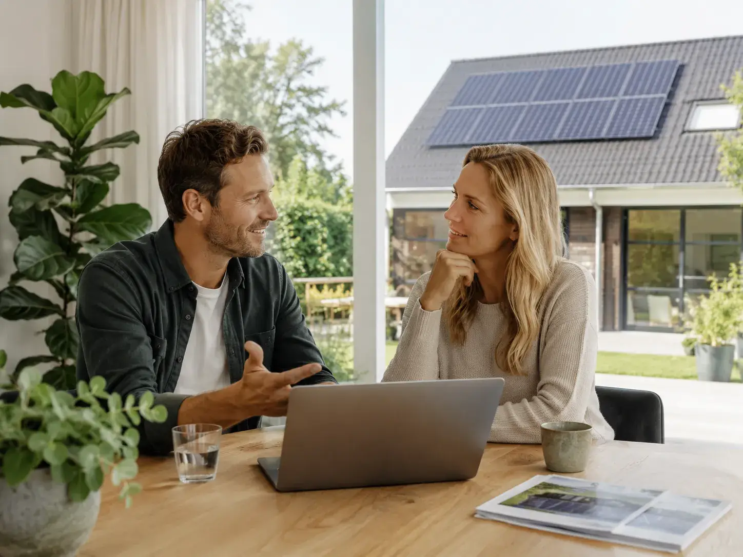 Zonnepanelenadviseur bespreekt vragen met een huiseigenaar aan tafel, met een woning met zonnepanelen op de achtergrond
